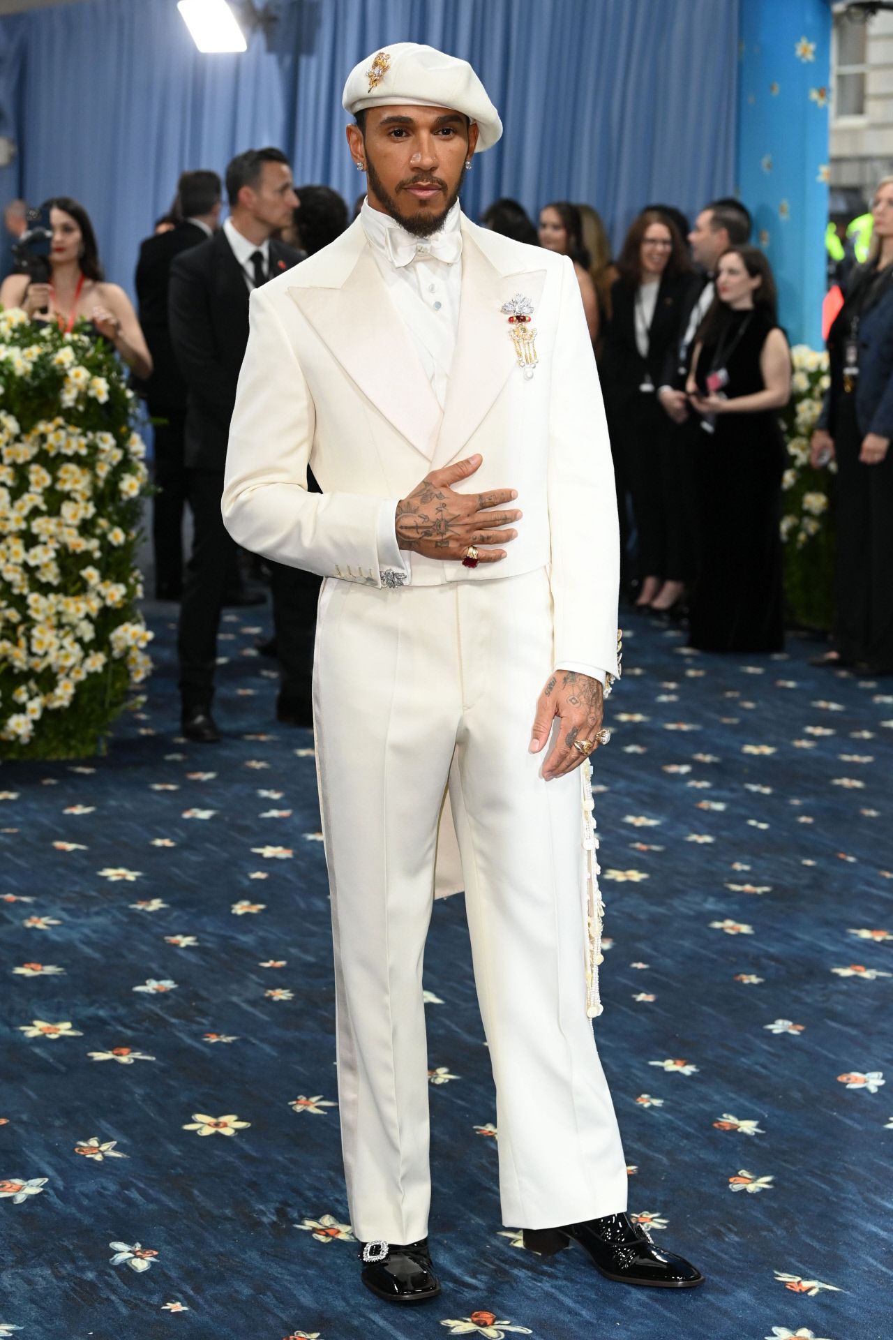 Lewis Hamilton, uno de los anfitriones este año, eligió un traje blanco de la diseñadora inglesa Grace Wales hecho a medida para la ocasión. Se trata de un conjunto que incluye pantalón recto, americana crop y abrigo. Lo combinó con boina y joyas vintage, incluido un llamativo broche que llevó en la solapa.